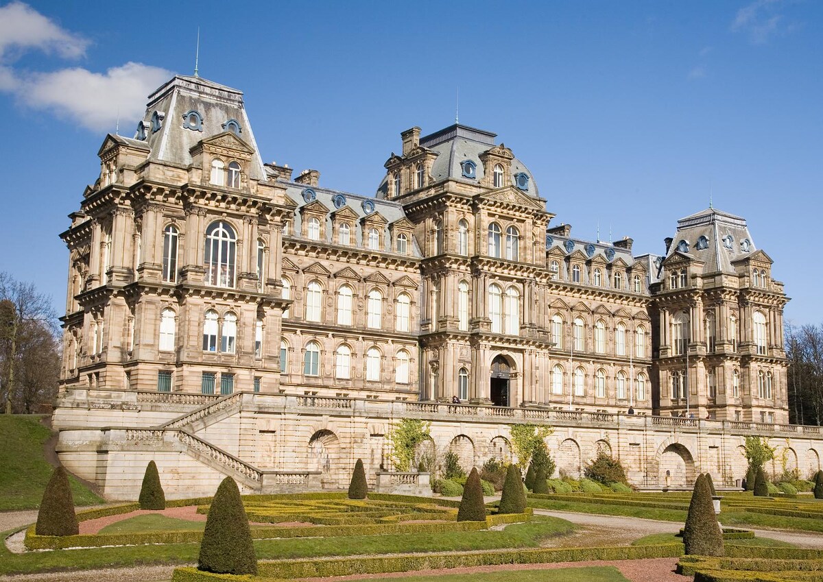 An impressive historical building is showcased, featuring grand architecture with multiple levels and ornate details. The exterior is adorned with large windows and decorative elements. Well-manicured gardens surround the structure, presenting neatly trimmed hedges and pathways leading to the entrance.