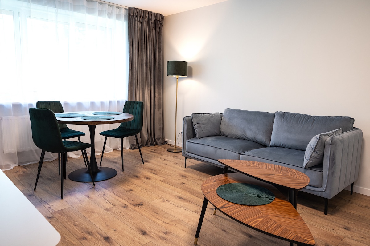 A comfortable living area features a grey sectional sofa beside a uniquely shaped wooden coffee table. A round dining table with green chairs is positioned near a large window, allowing natural light to brighten the space. Soft drapes complement the decor.