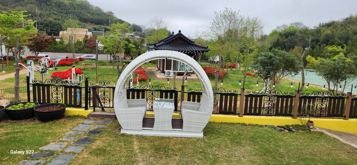 A white wicker swing is positioned in the foreground, surrounded by a lush green lawn. A traditional Korean house is visible in the background, framed by vibrant greenery and colorful flower beds, creating a serene outdoor space for relaxation.