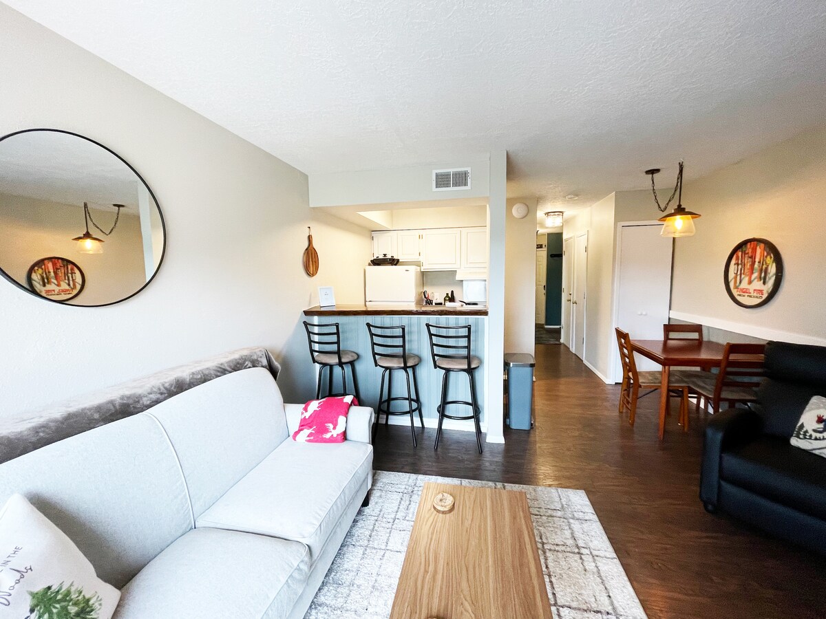 A modern living area is furnished with a light gray couch, a wooden coffee table, and a round mirror. A kitchen bar with three stools is visible in the background, alongside a dining table with four chairs. Light-colored walls and hardwood flooring create a cohesive look.