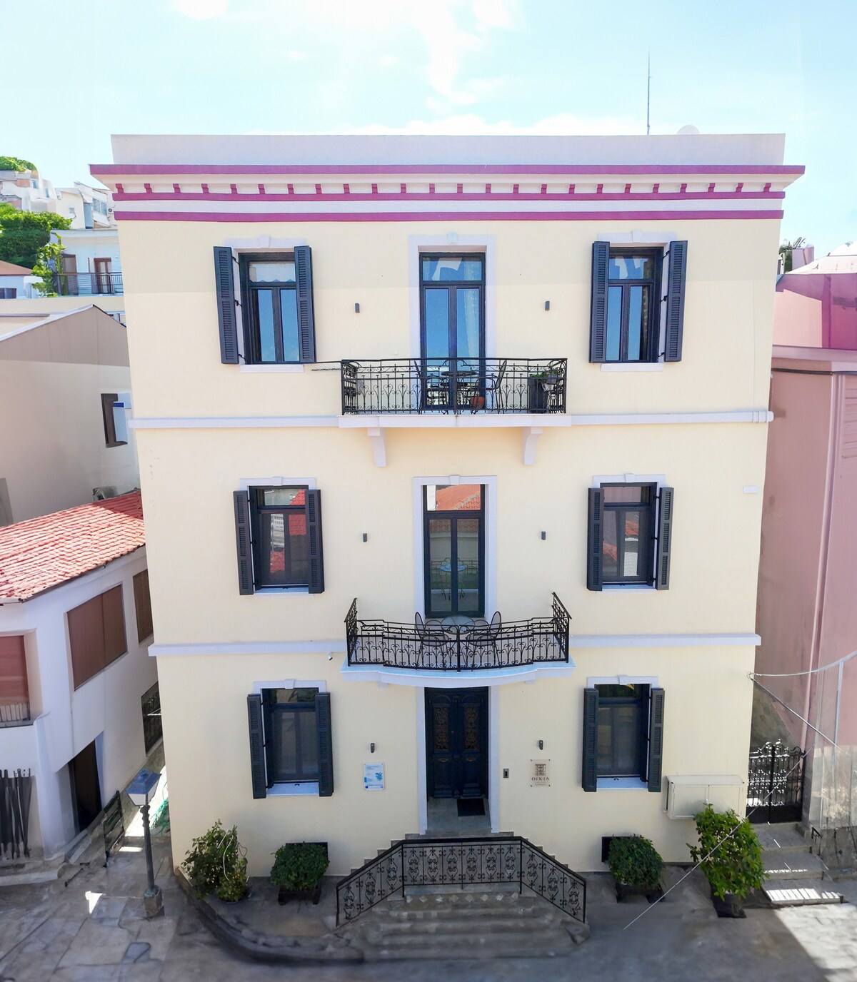 The elegant neoclassical building showcases a light-colored facade with symmetrical balconies adorned with black wrought-iron railings. Large, dark window shutters contrast with the exterior, while meticulously arranged plants flank the entrance, enhancing the inviting appearance of the structure.