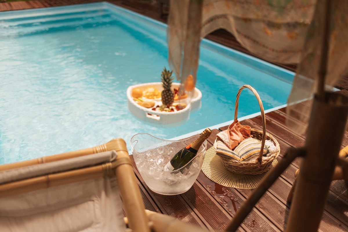 A refreshing private pool is shown, surrounded by wooden decking. A cooler filled with ice and a bottle of champagne rests beside a woven basket filled with assorted fruits. Nearby, a soft blanket adds comfort, suggesting a relaxed atmosphere for outdoor enjoyment.