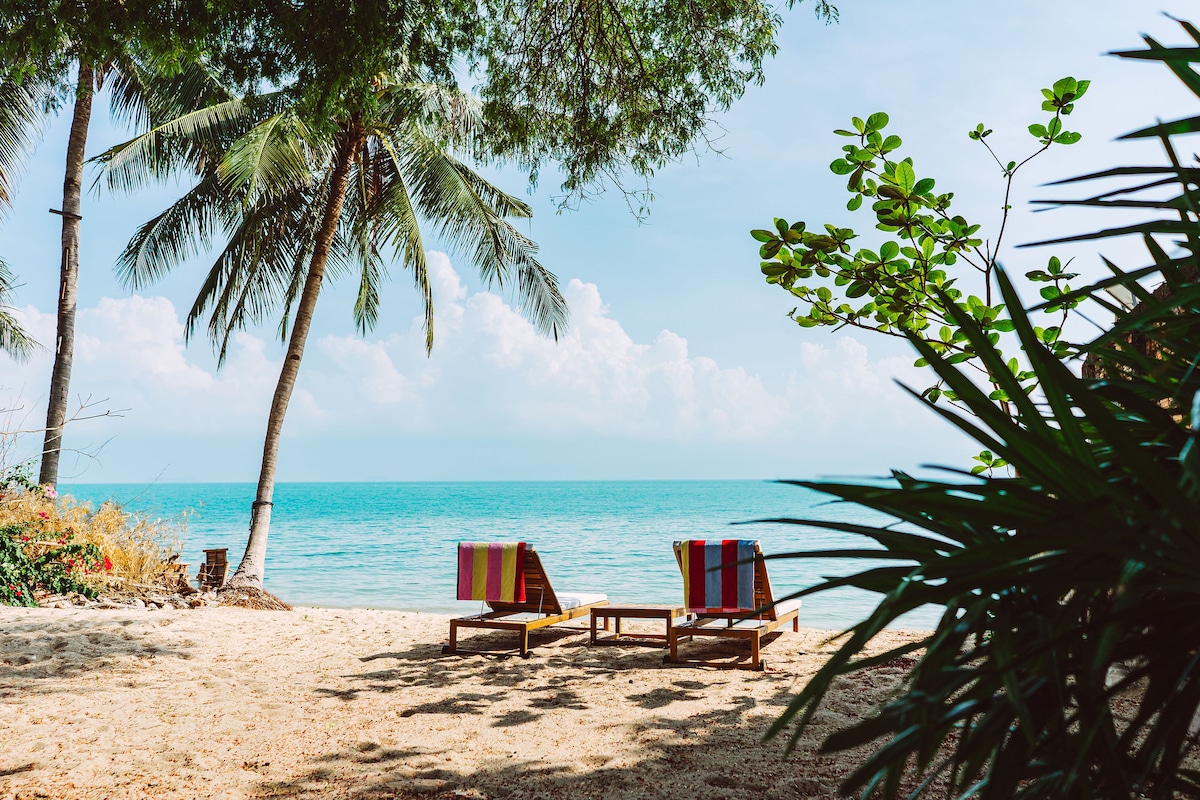 Two sunbeds with colorful striped towels are arranged on sandy beachfront. Palm trees offer shade, and lush greenery is visible in the foreground. The turquoise sea stretches out to the horizon beneath a clear sky, inviting relaxation and tranquility.