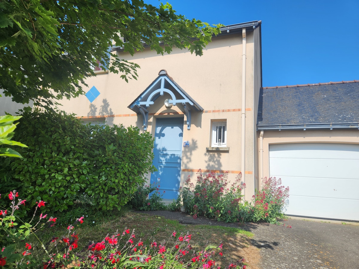 The exterior of a charming house is visible, featuring a blue front door framed by greenery and colorful flowers. A garage is positioned to the right, alongside a clear pathway leading to the entrance. Sunlight illuminates the façade, creating a welcoming appearance.