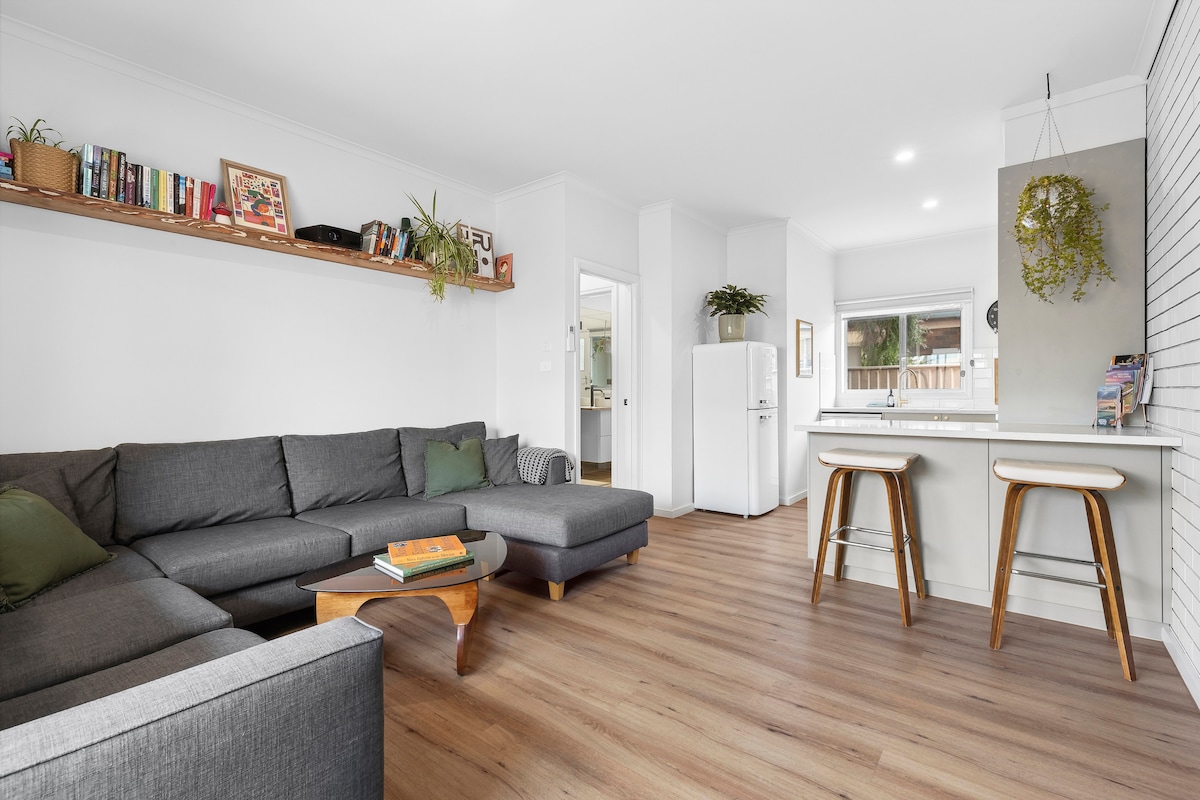 A spacious living area is highlighted by a grey sectional sofa accented with green cushions. A wooden coffee table sits at the center, while a bookshelf displays various books on the wall. A bright kitchen area is visible in the background, featuring two bar stools.