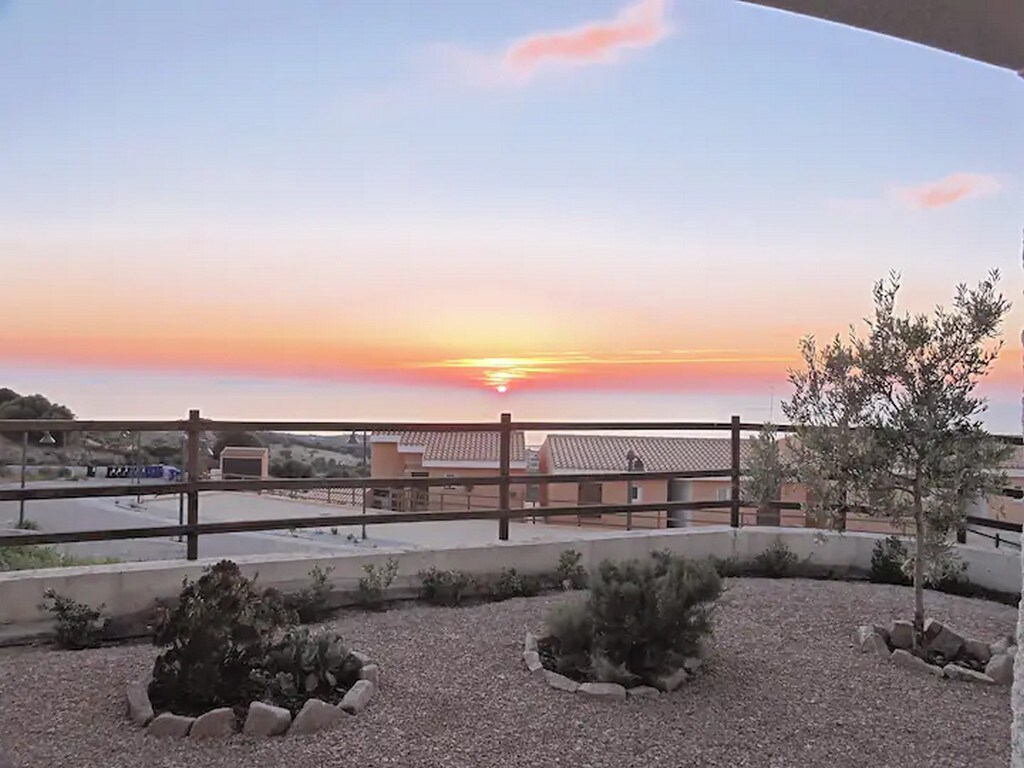 A sunset view is presented over the horizon, with warm orange and pink hues reflecting on the sky. The foreground features a landscaped area with small shrubs and decorative stones, and a railing offers a clear perspective of the surrounding scenery.