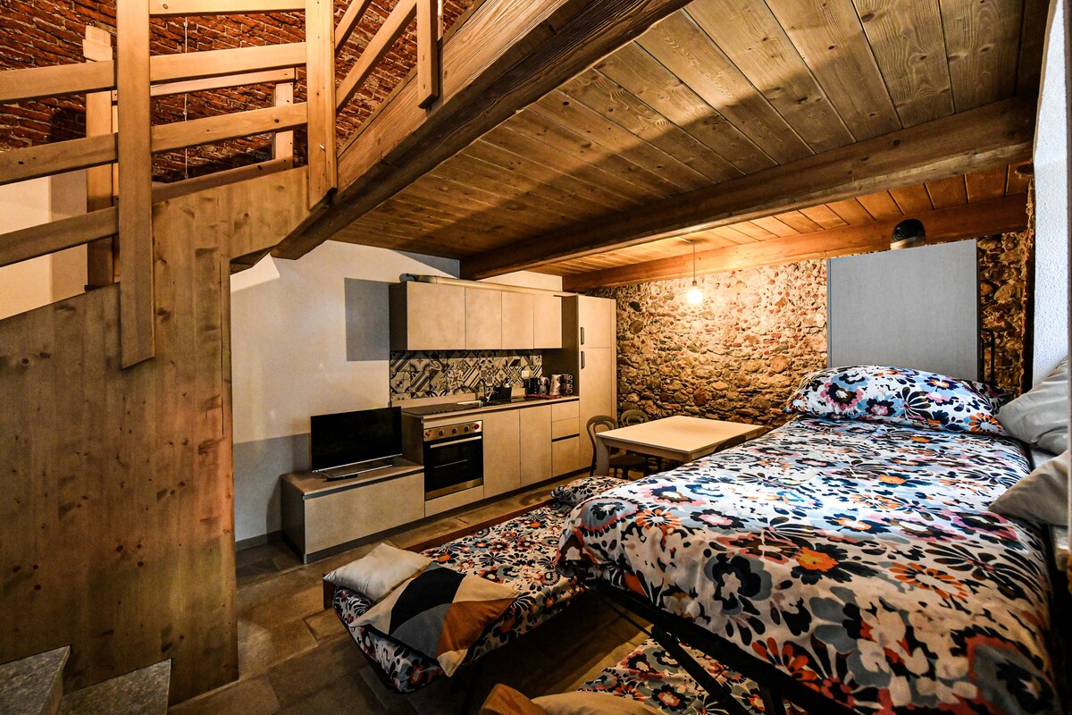 A spacious studio is highlighted with natural wood beams and a light-colored stone wall. A large bed with a floral-patterned cover occupies one side, while a kitchenette with modern appliances is visible. A small dining table and chairs provide a functional space beneath the lofted area.