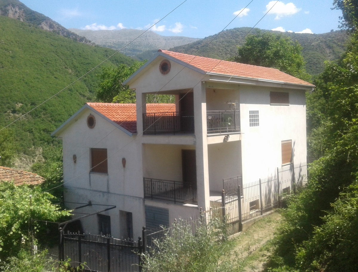 A two-story house is situated amidst lush greenery and rolling hills. The building features a red-tiled roof, balconies on both levels, and a fenced yard. Surrounding trees enhance the natural setting, while the distant mountains provide a scenic backdrop.