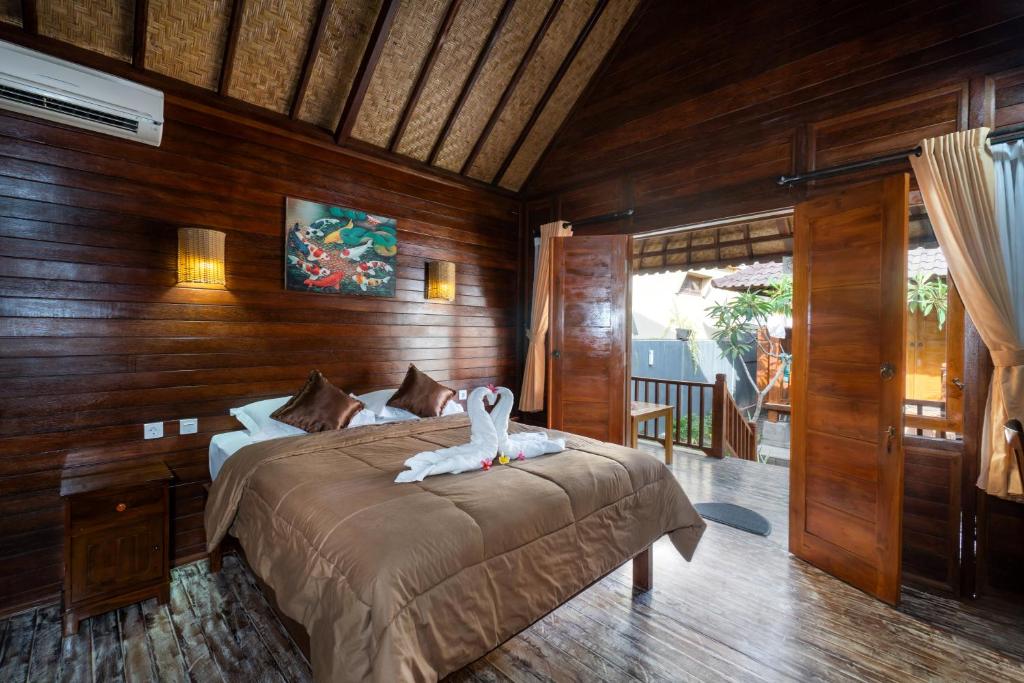 A wooden interior features a spacious bed adorned with a decorative arrangement of towels. Warm lighting is provided by wall sconces, and large doors open to a terrace, offering views of the outdoor area. Natural textures are highlighted in the ceiling and walls.