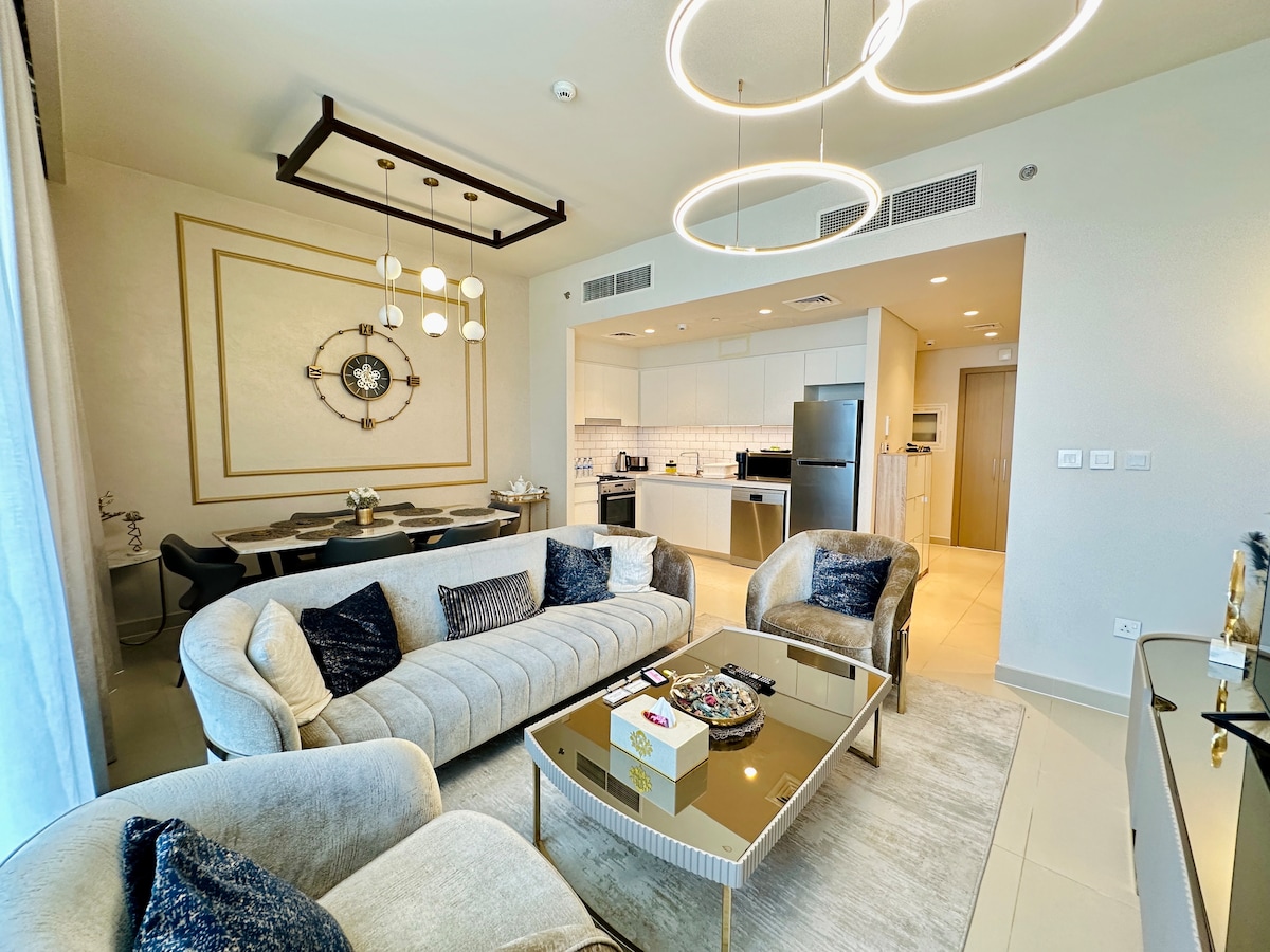 A stylish living area features a plush sofa and elegant armchairs surrounding a coffee table. A modern chandelier hangs above, and a dining space with a large table is visible in the background. The kitchen, equipped with built-in appliances, is seamlessly integrated into the open-concept layout.