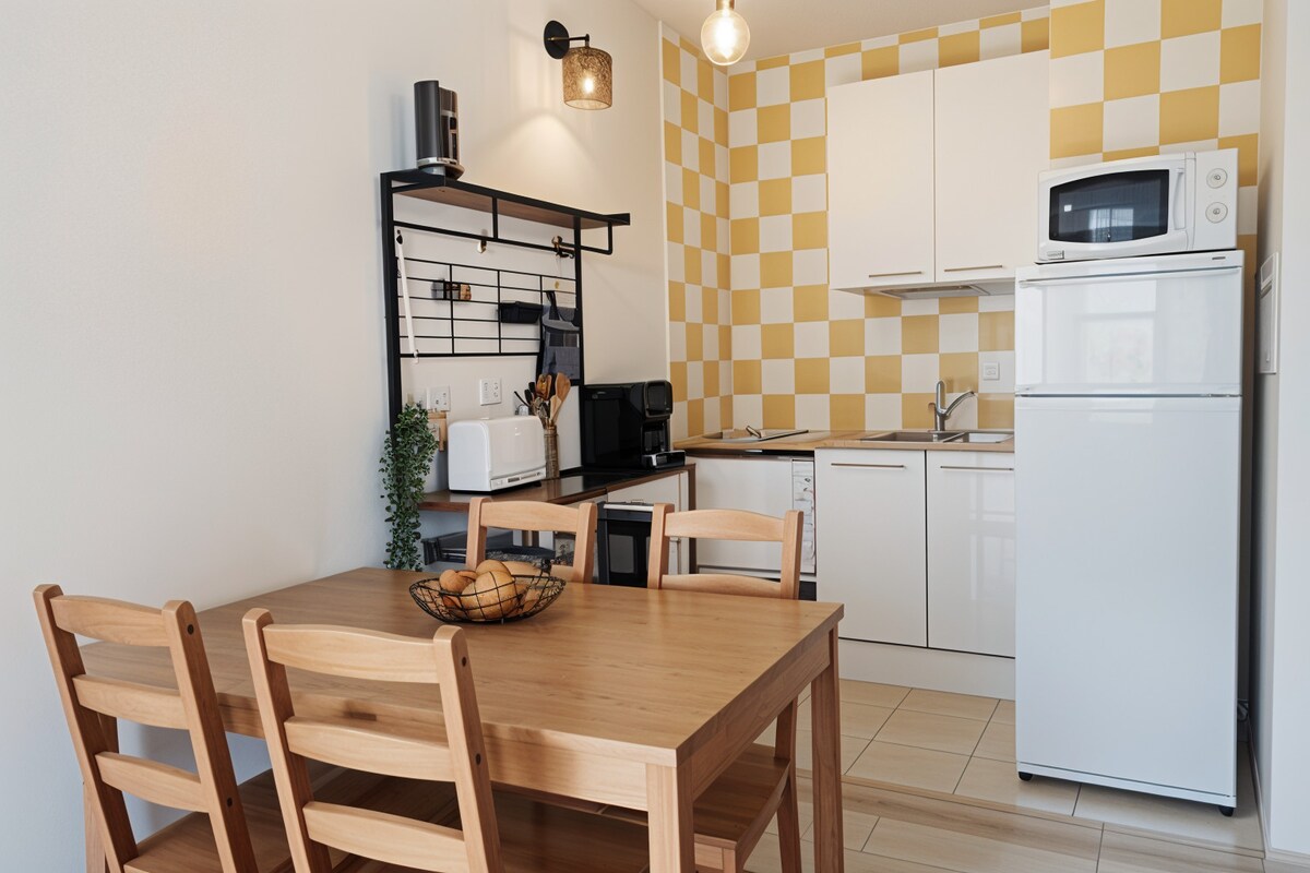 A bright kitchen space is seen, featuring a compact dining table with four wooden chairs. Yellow-checkered walls complement the white cabinetry, which holds a microwave and a refrigerator. Small appliances and cookware are organized on shelving, creating an inviting cooking area.