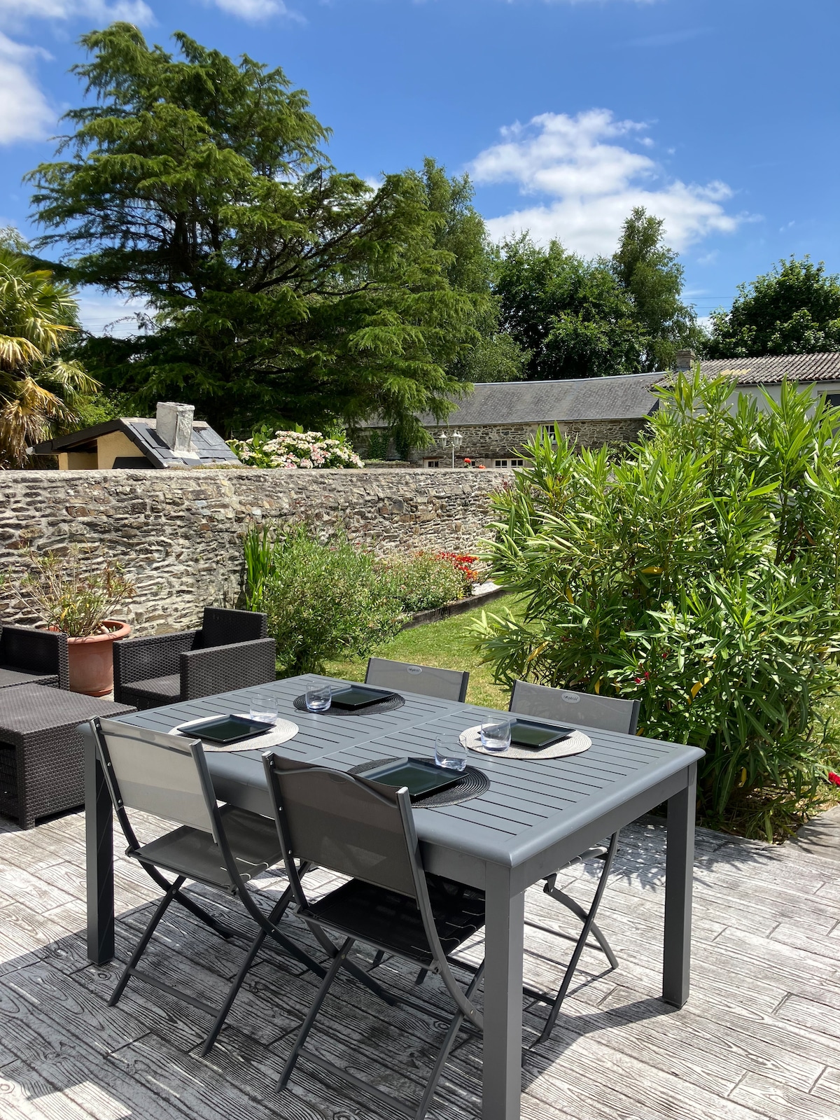 An outdoor dining area features a gray table set for four, accompanied by plates and utensils. Lush greenery surrounds the space, with a stone wall in the background and a sunny sky overhead. Comfortable seating is positioned nearby, inviting relaxation.