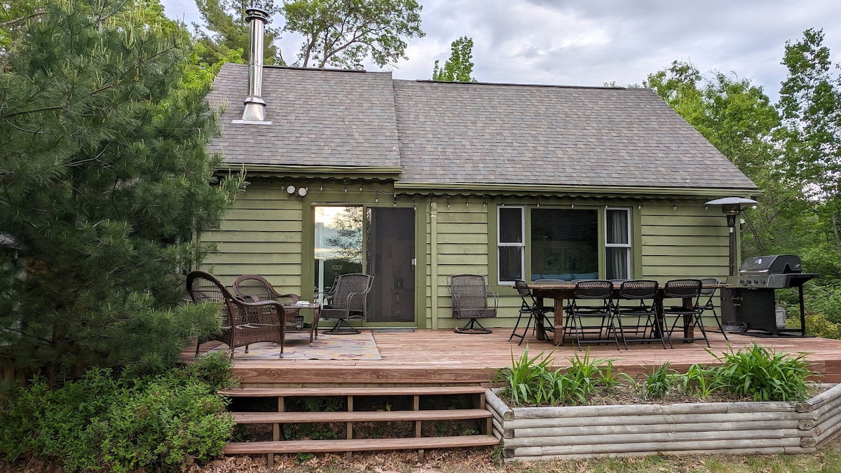 The cabin features a rustic green exterior, complemented by a spacious wooden deck. The deck is furnished with a dining table and chairs, along with a grill. Large windows enhance natural light and views of the surrounding greenery.