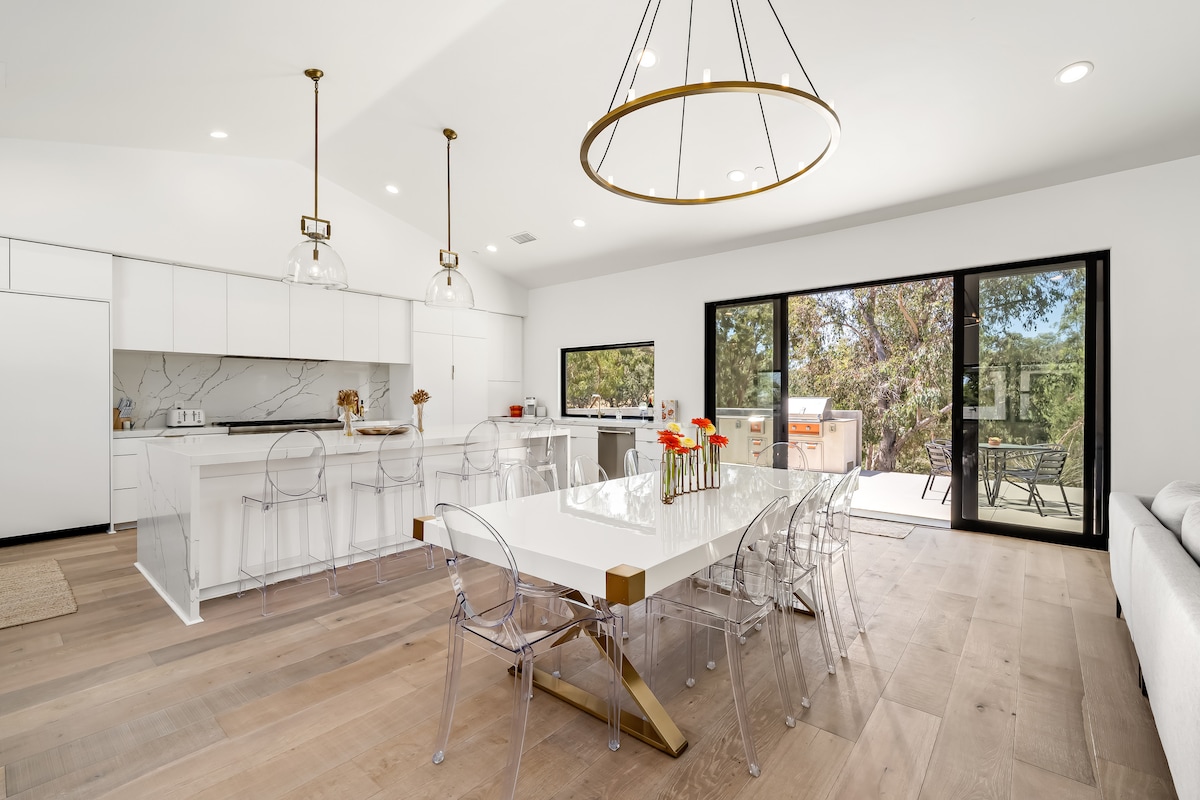 An open-plan kitchen and dining area features a modern white table surrounded by transparent chairs. The kitchen showcases sleek cabinetry and marble accents. Tall glass doors invite natural light and provide a view of the outdoor space, enhancing the connection to the surrounding landscape.