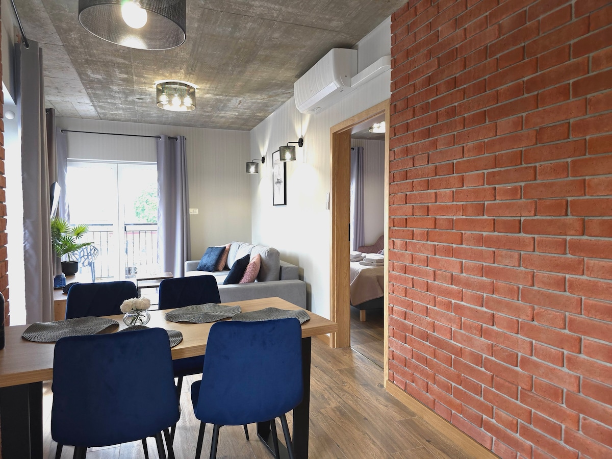 A dining area is visible, featuring a wooden table surrounded by blue chairs. Soft gray tones and natural light create a calm atmosphere. A cozy seating area with a gray sofa is positioned to the side, complemented by a view of a balcony through large glass doors.