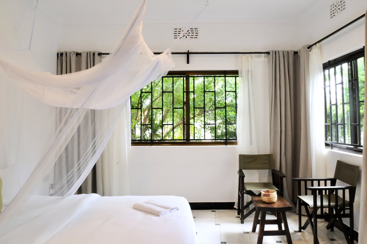 The tranquil bedroom features a spacious bed adorned with fresh linens, complemented by a mosquito net overhead. A wooden table and two chairs offer a cozy sitting area, while large windows with sheer curtains invite natural light and provide garden views.
