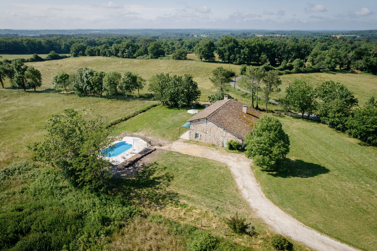 An aerial view showcases a traditional house surrounded by lush greenery and open fields. A secure swimming pool is visible beside the house, offering a private outdoor space. The property is bordered by trees, enhancing the natural setting.