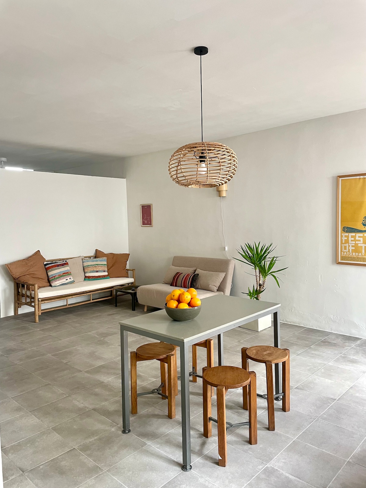 A spacious living and dining area is presented, featuring a simple table surrounded by three wooden stools. A bowl of oranges is positioned at the center. A sofa with decorative cushions and a potted plant add a touch of comfort to the neutral-toned space.
