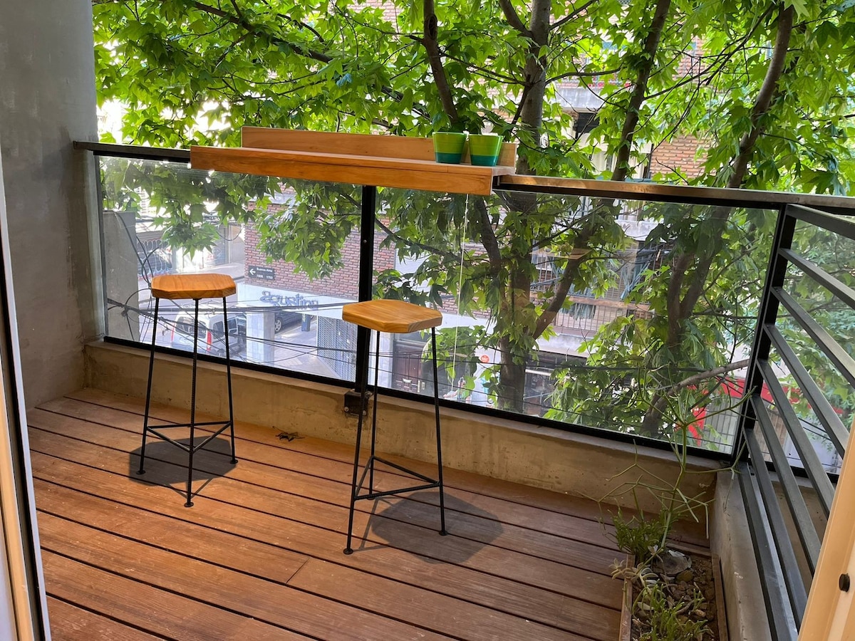 A balcony area is presented with two wooden stools positioned next to a low wooden table. Lush greenery is visible through the glass railing, creating a natural backdrop. The flooring features wooden planks, enhancing the outdoor ambiance.