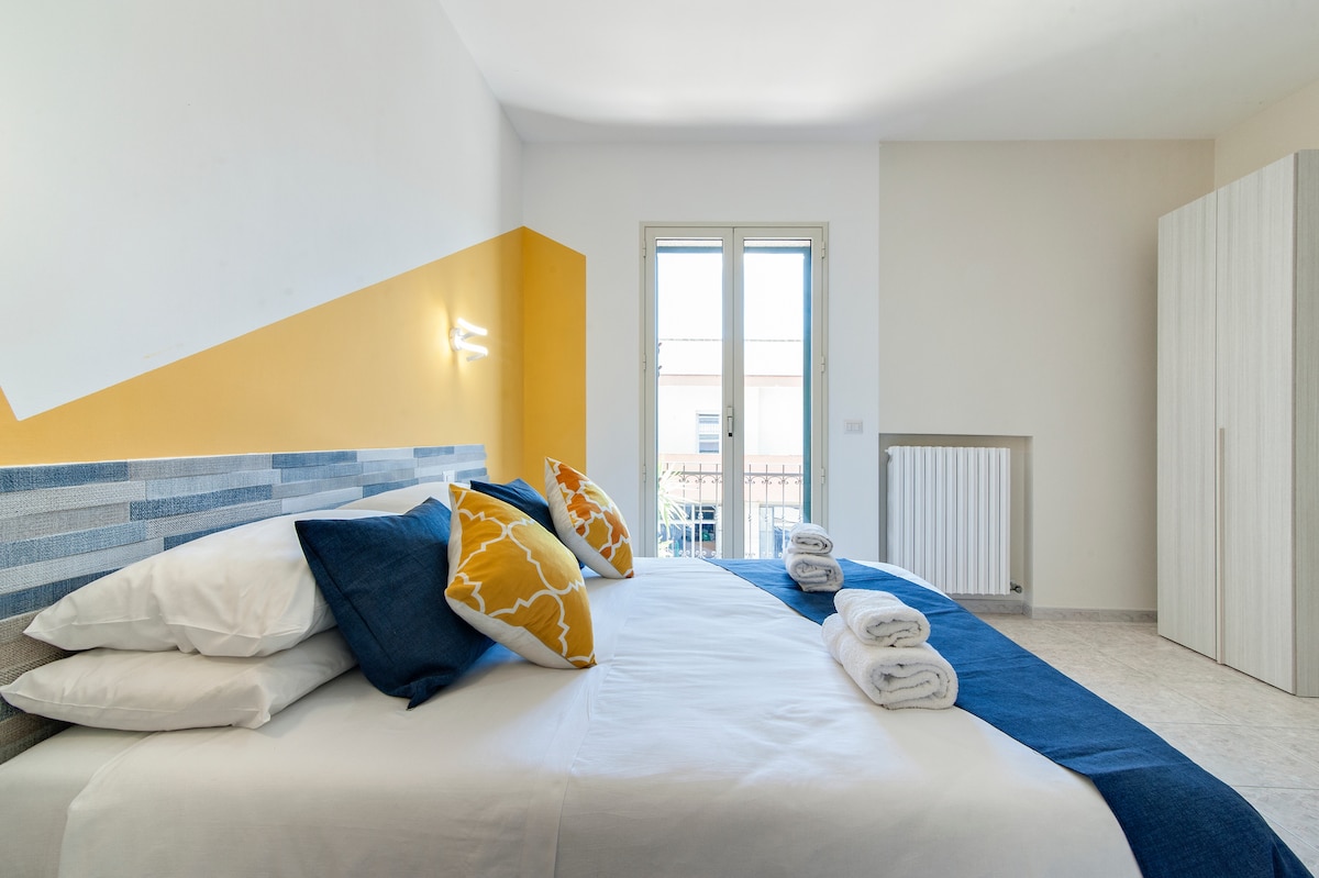 A well-lit bedroom features a spacious bed with white linens and decorative pillows. Two windows allow natural light, while a wardrobe and radiator are seen in the corner. Bright yellow accents complement the decor, creating a cheerful atmosphere.