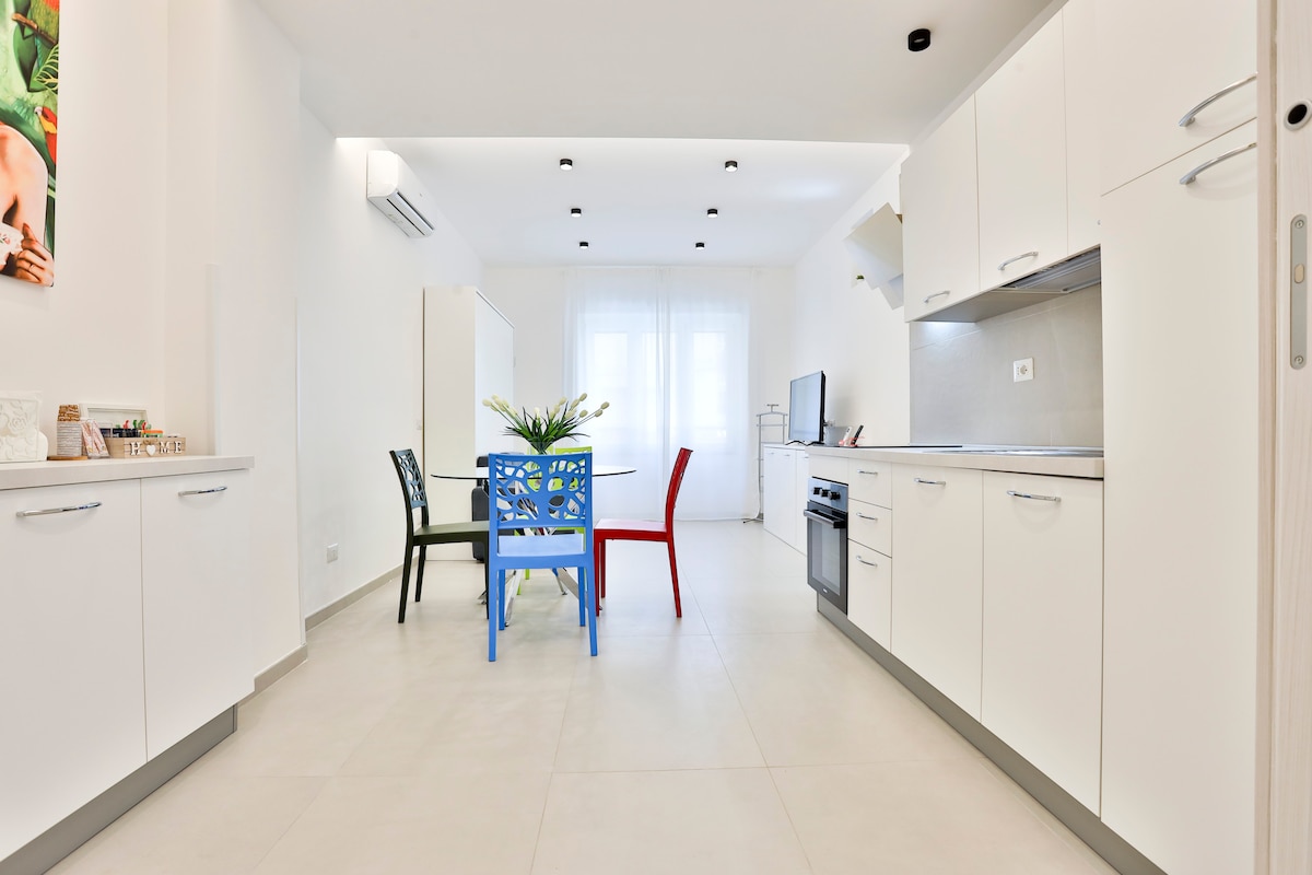 The bright kitchen area features modern white cabinetry and a sleek countertop. A small dining table is surrounded by colorful chairs, including a blue one, offering a cheerful dining space. Natural light floods the room through a large window, enhancing the airy feel.