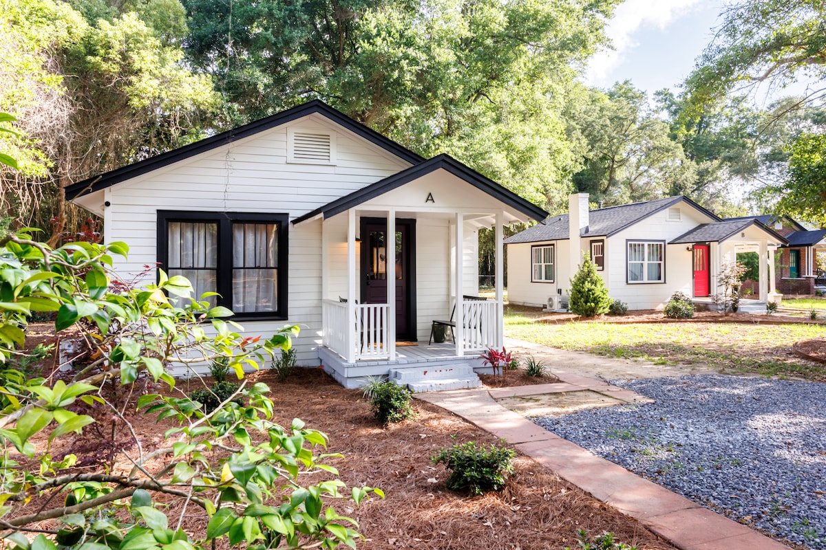 A charming villa exterior is shown, featuring a white facade with black accents. A porch with steps invites entry, bordered by colorful landscaping and gravel paths. Adjacent villas are visible, surrounded by lush greenery, conveying a serene and welcoming atmosphere.