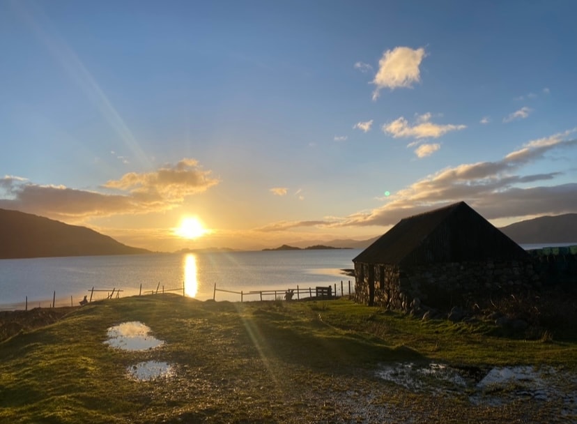 A serene landscape captures the sunset over a calm body of water, with reflections shimmering on the surface. A rustic building stands nearby, surrounded by grassy grounds and patches of water. The distant hills frame the scene, enhancing the natural beauty.