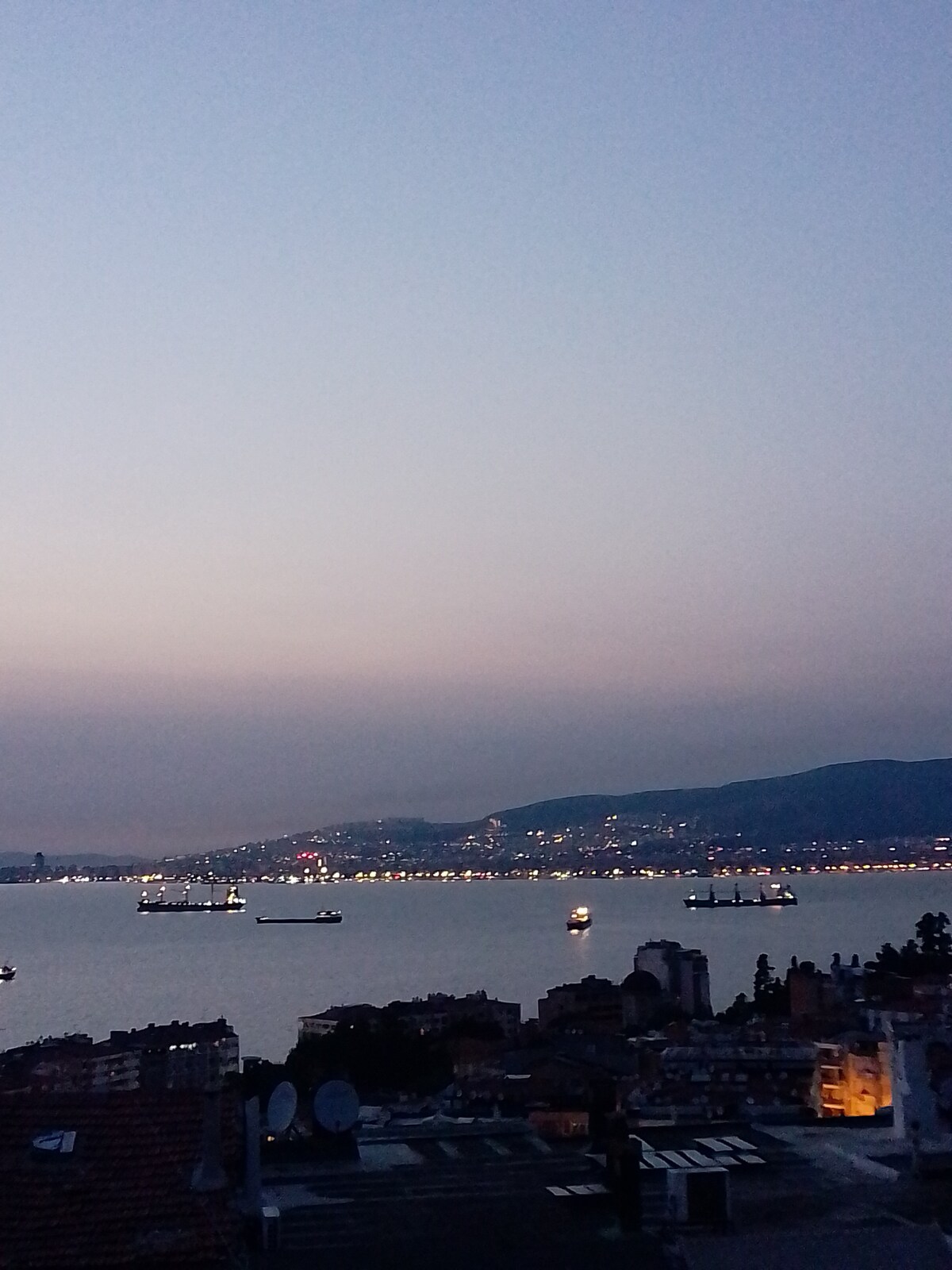A coastal view is captured during twilight, revealing the silhouette of ships anchored in the water. City lights glimmer in the distance along the shoreline, with a gentle gradient of blue and pink hues in the sky.