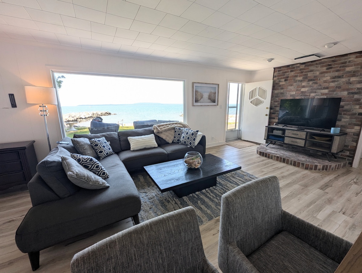 A spacious living area features a large sectional sofa adorned with decorative pillows, facing a modern television. Large windows showcase views of the waterfront, while a coffee table sits centrally on the light-toned floor. A stone accent wall adds texture to the room.