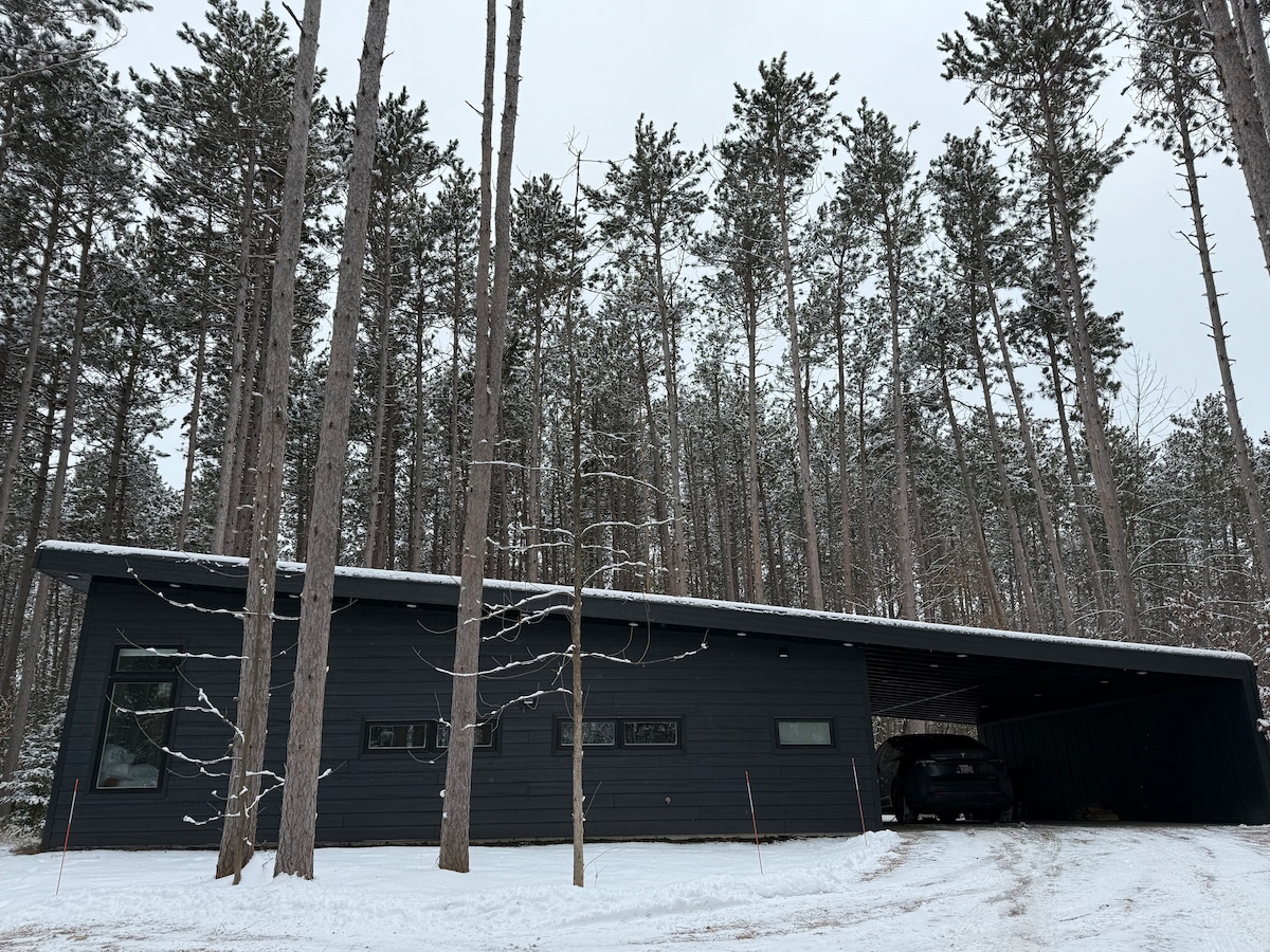 The exterior of a modern home is framed by tall pine trees, dusted with snow. A car is parked under an attached carport on one side, while large windows provide a glimpse into the interior. The sleek design complements the serene wooded environment.