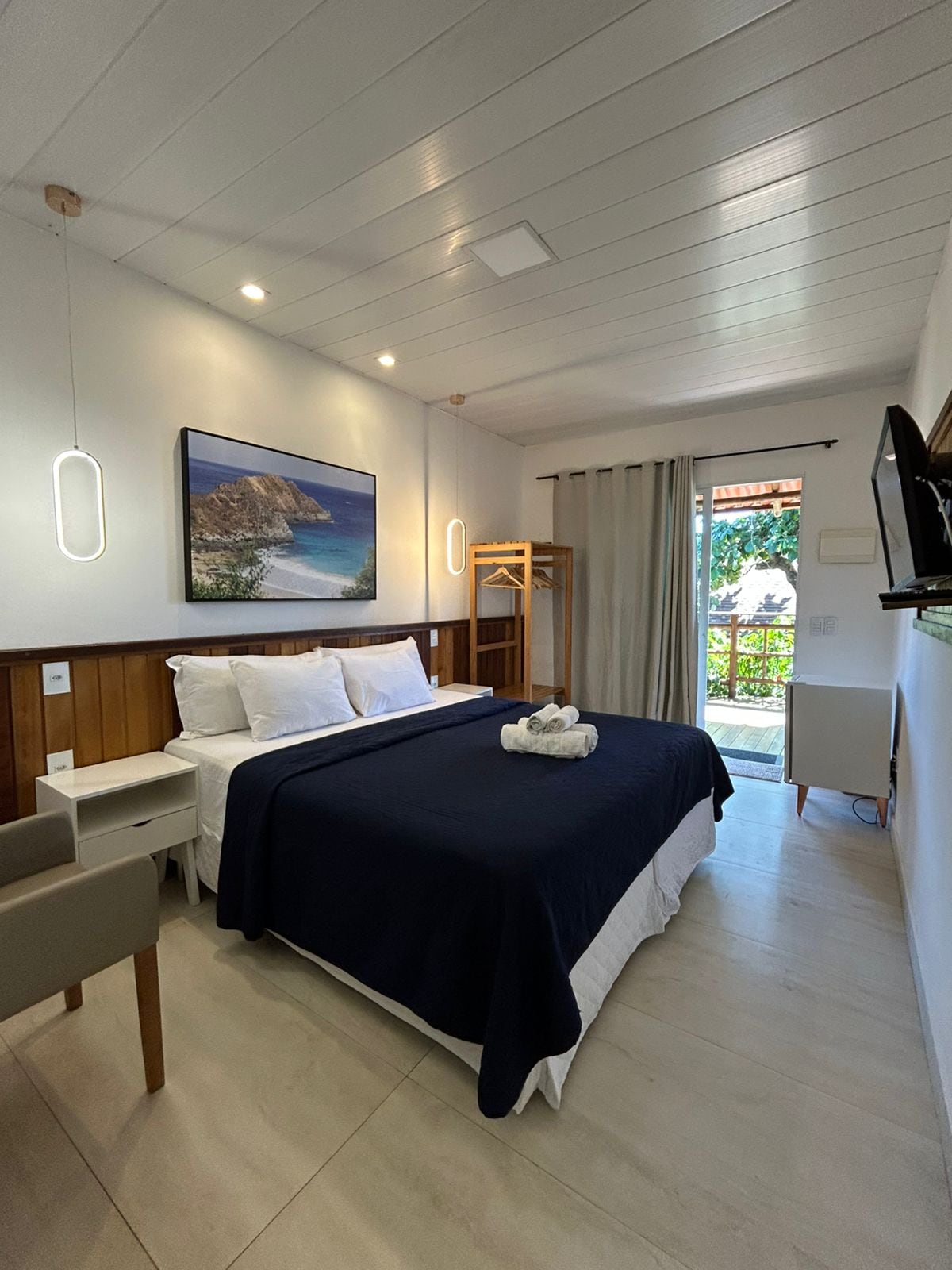 A serene guest room is highlighted by a spacious bed with dark bedding. A flat-screen TV is mounted on the wall, and a wooden closet is visible. Natural light enters through sliding glass doors, revealing a view of the outdoor area with greenery.