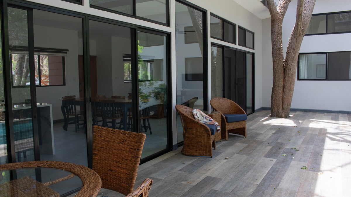 A spacious terrace is adorned with comfortable seating arrangements, including two woven chairs and a small table. Large glass doors enhance natural light and provide views of the surrounding trees and the interior living area.