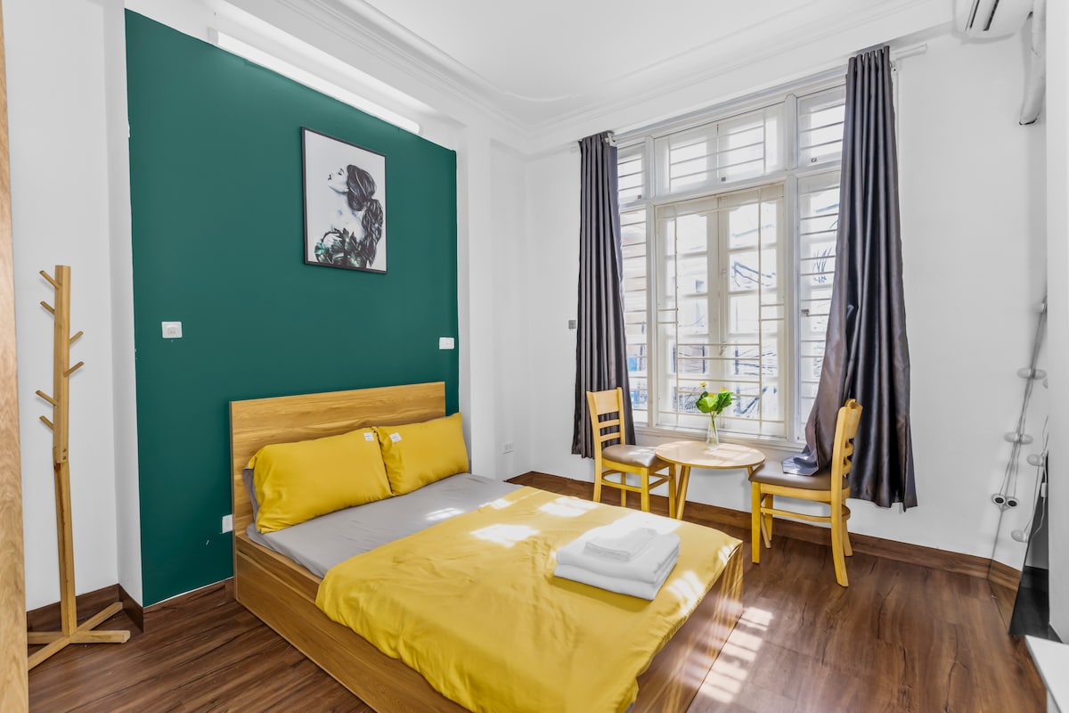 A small room features a wooden bed with yellow bedding, complemented by a simplistic wall art piece. A round table and two chairs sit beside a large window, allowing natural light to fill the space. Curtains frame the window, and a coat rack stands nearby.