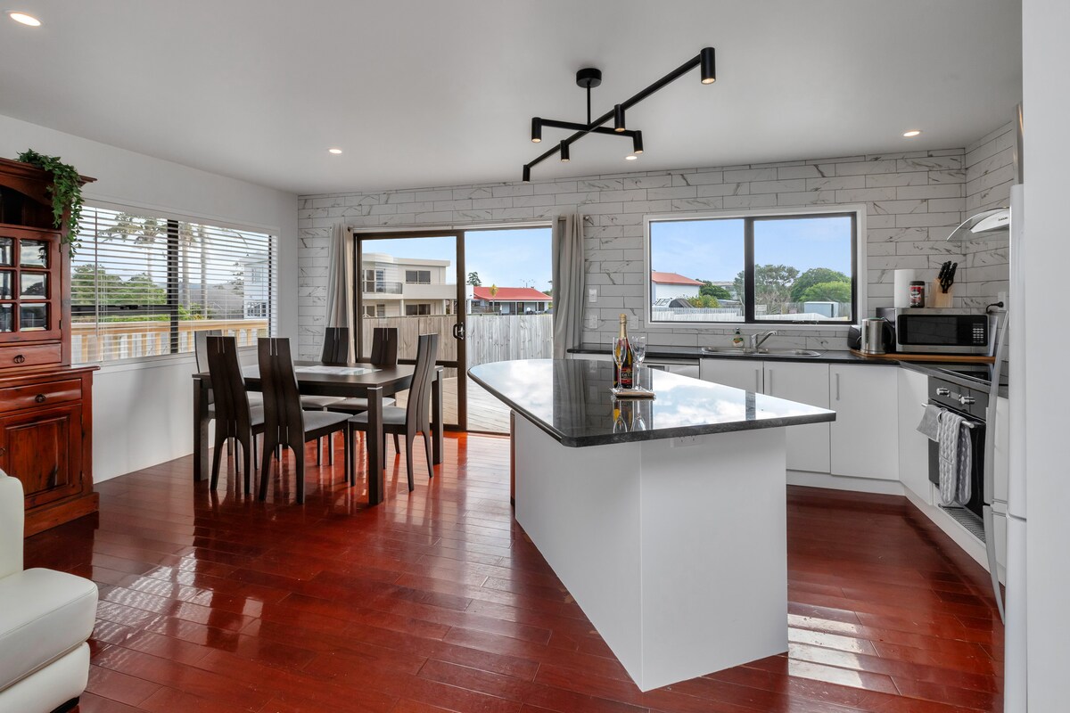An open-plan kitchen and dining area features modern fixtures and a large island with seating. Natural light floods the space through multiple windows, illuminating the warm wooden floors. A dining table accommodates several guests, creating an inviting atmosphere for meals and gatherings.