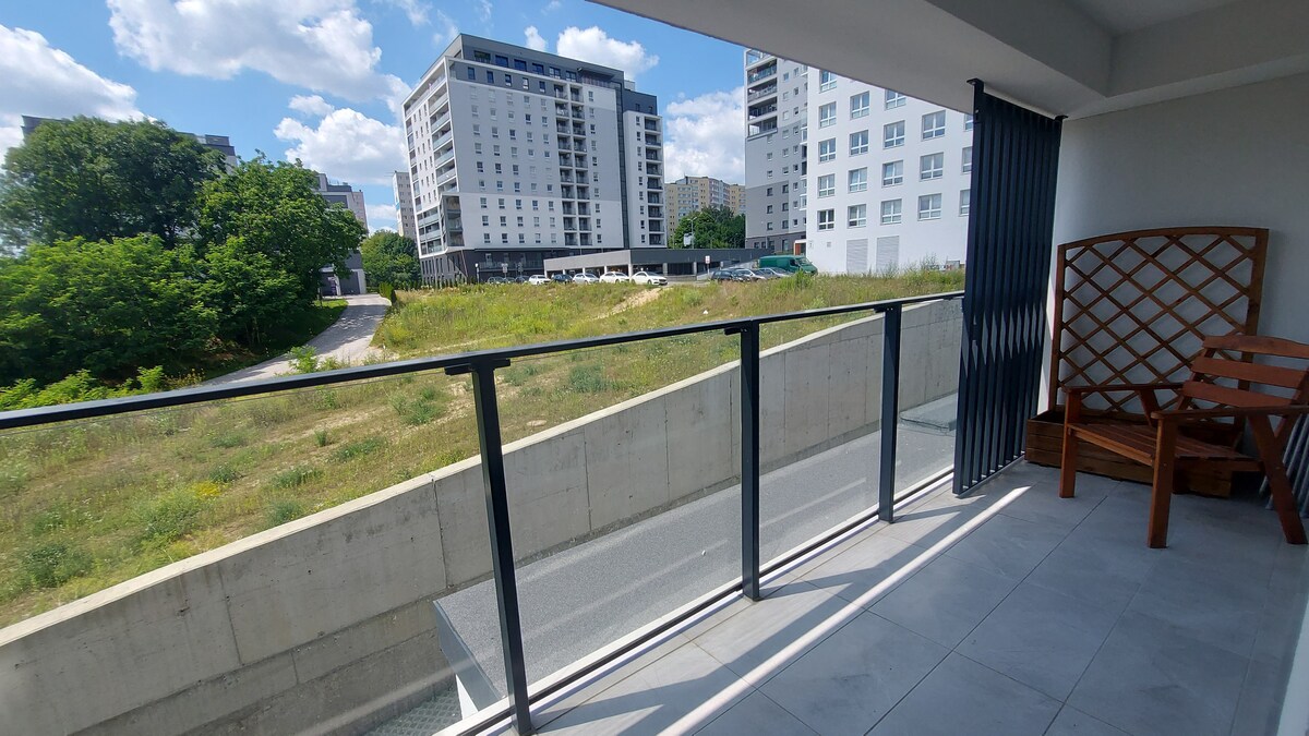 A spacious balcony features a sturdy wooden bench and a railing overlooking a natural area with greenery. The adjacent buildings are visible in the background, enhancing the view. Bright sunlight illuminates the tiled floor, contributing to an open atmosphere.