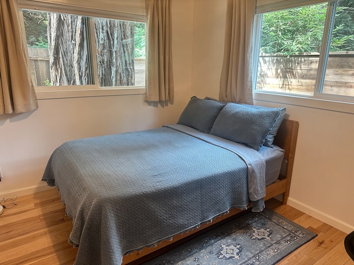 The main-level third bedroom offers a full-sized bed and an office setup with a standing desk, monitor, wireless keyboard, and mouse. Two windows bring in wonderful afternoon light, creating a perfect blend of comfort and productivity.