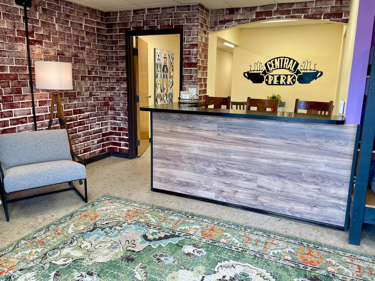 A welcoming reception area is highlighted by a dark wooden counter in front of a vibrant brick wall, featuring the text "CENTRAL PERK". A cozy gray armchair sits adjacent to the counter, alongside a patterned area rug that adds a touch of color to the space.