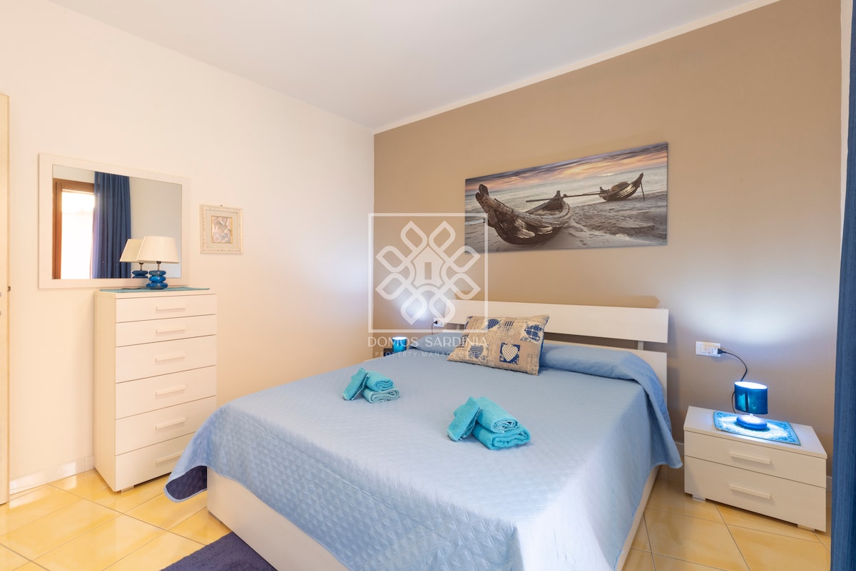 A spacious bedroom is furnished with a comfortable bed dressed in light blue linens. A nightstand with a lamp is situated beside the bed, complemented by a dresser across from it. Sunlight filters through a window, illuminating the serene artwork on the wall.