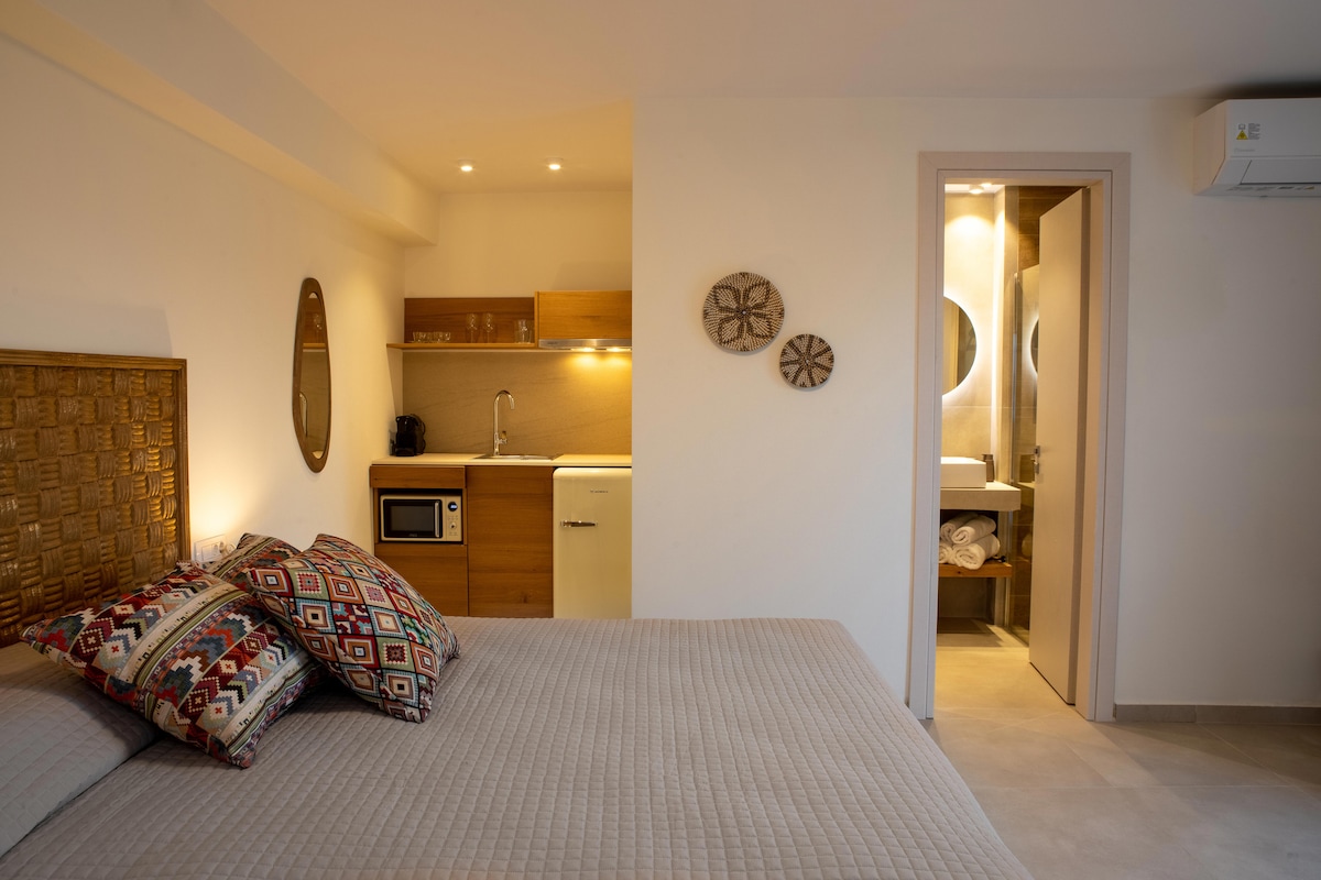 A bright double room features a spacious bed with decorative pillows. A kitchenette equipped with a microwave and refrigerator is visible in the background. The private bathroom entrance is positioned to the right, while soft lighting enhances the overall calmness of the space.