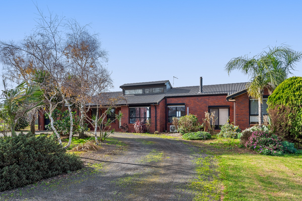 The large brick home features a welcoming facade surrounded by lush greenery and landscaped gardens. A driveway leads to the entrance, with palm trees and various shrubs enhancing the outdoor aesthetic. A well-maintained lawn adds to the inviting appearance of the property.