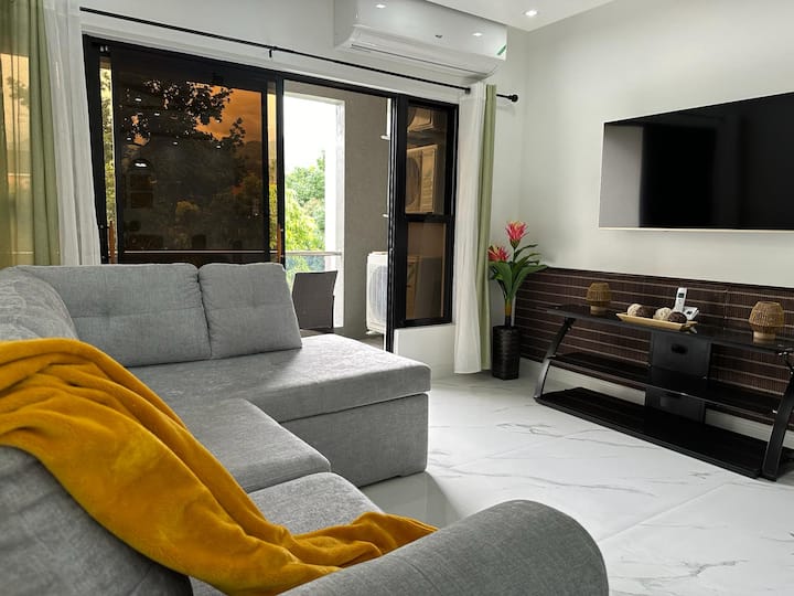 Sebule ina sofa ya kijivu na televisheni ya skrini bapa iliyowekwa ukutani, na meza nyeusi ya kifahari hapa chini. Ua la rangi ya waridi na kijani huongeza mguso wa rangi kwenye kona na milango ya kuteleza iliyo wazi kidogo hutoa mwonekano wa roshani ya nje.