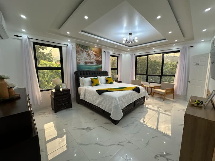 Chumba cha kulala cha msingi kilicho na kitanda chenye bendera ya Jamaika na vigae vya marumaru. Meza mbili kando ya kitanda zilizo na taa zinakamilisha sehemu hiyo. Chumba hicho kina sehemu ya kukaa ya watu wawili iliyo na meza ndogo, madirisha pande zote mbili za kitanda na televisheni ukutani.