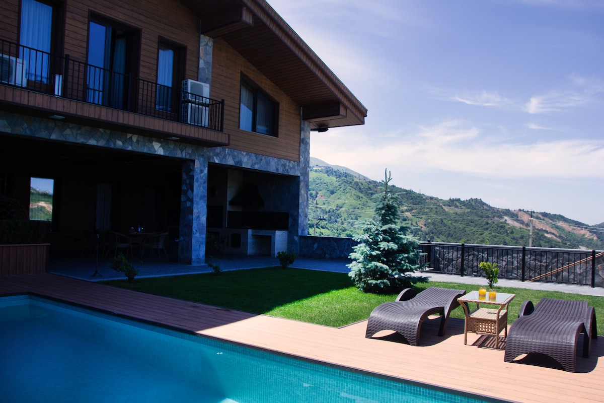 An outdoor area features a swimming pool surrounded by wooden decking. Two lounge chairs face the pool, accompanied by a small table. The house showcases a stone facade with large windows and an outdoor grill area, framed by lush greenery and mountain views.