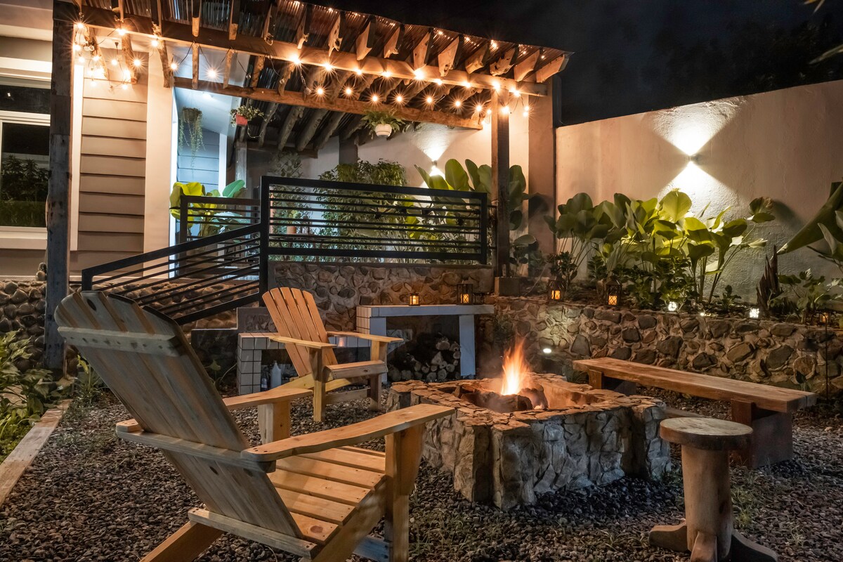 An outdoor bonfire area is highlighted by a circular stone seating arrangement surrounding a warm fire pit. Wooden chairs provide comfortable seating under soft lighting, with lush greenery and decorative stones enhancing the space. The area is set against a backdrop of a well-lit villa.