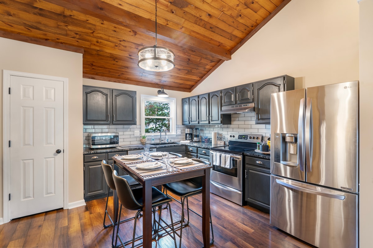 A modern kitchen features dark cabinetry and stainless steel appliances. A wooden ceiling adds warmth to the space. A dining table is set for four, placed near a window that offers natural light. The countertop is equipped with essential kitchenware.