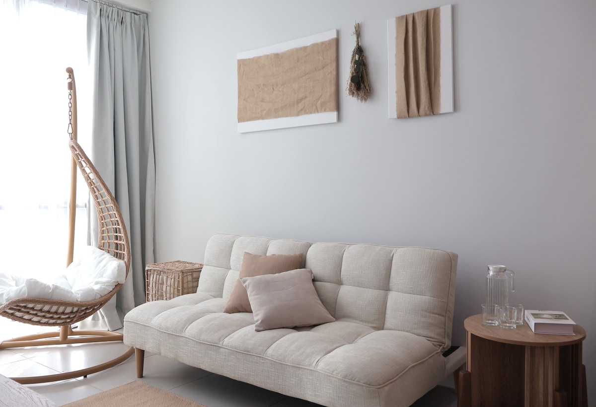 A light-filled living area features a comfortable sofa with neutral upholstery. A round wooden side table and a woven basket add texture. Two pieces of wall art in soft tones hang above the sofa, while a hanging chair in the corner offers a cozy seating option.