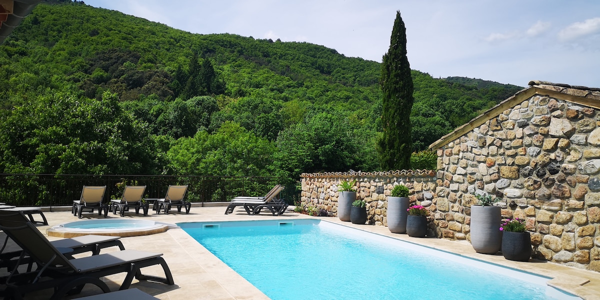 A shared swimming pool is surrounded by sun loungers, set against a backdrop of lush green hills. The area is adorned with planters featuring vibrant flowers, and a stone wall provides a rustic touch to the tranquil outdoor space.
