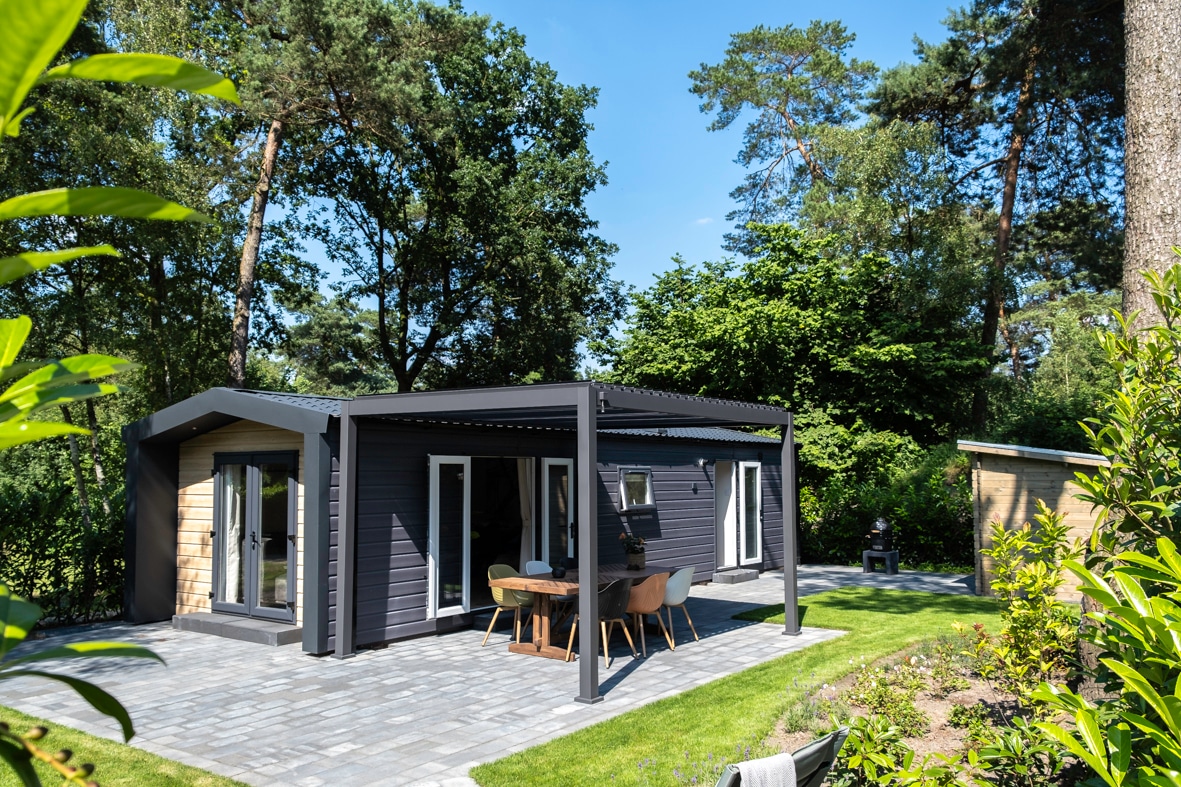 A modern chalet is displayed, featuring a spacious terrace with outdoor dining furniture. Large glass doors provide views into the interior, surrounded by a lush green landscape with trees and shrubbery.