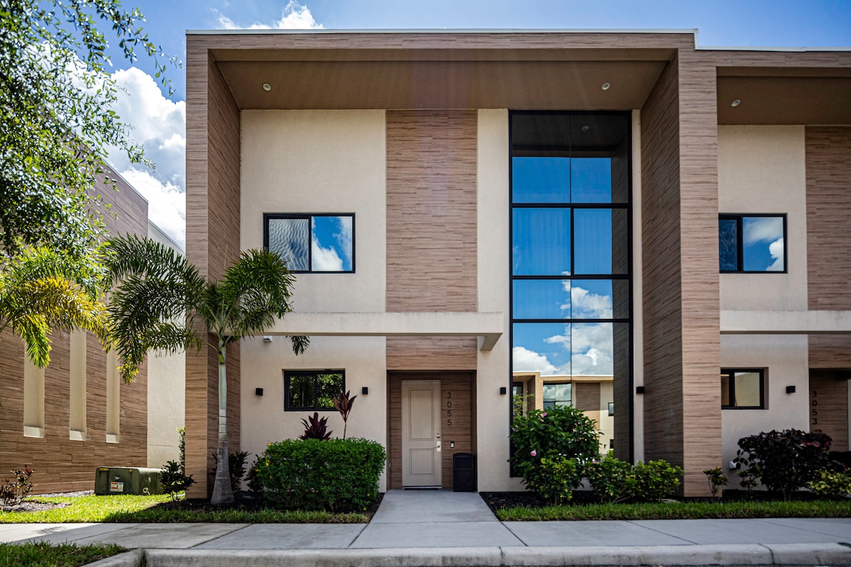 The modern two-story townhouse features a sleek facade with large glass windows reflecting the sky. Lush greenery and well-maintained landscaping frame the entrance, creating an inviting approach. The exterior showcases a blend of natural tones and minimalist design elements.