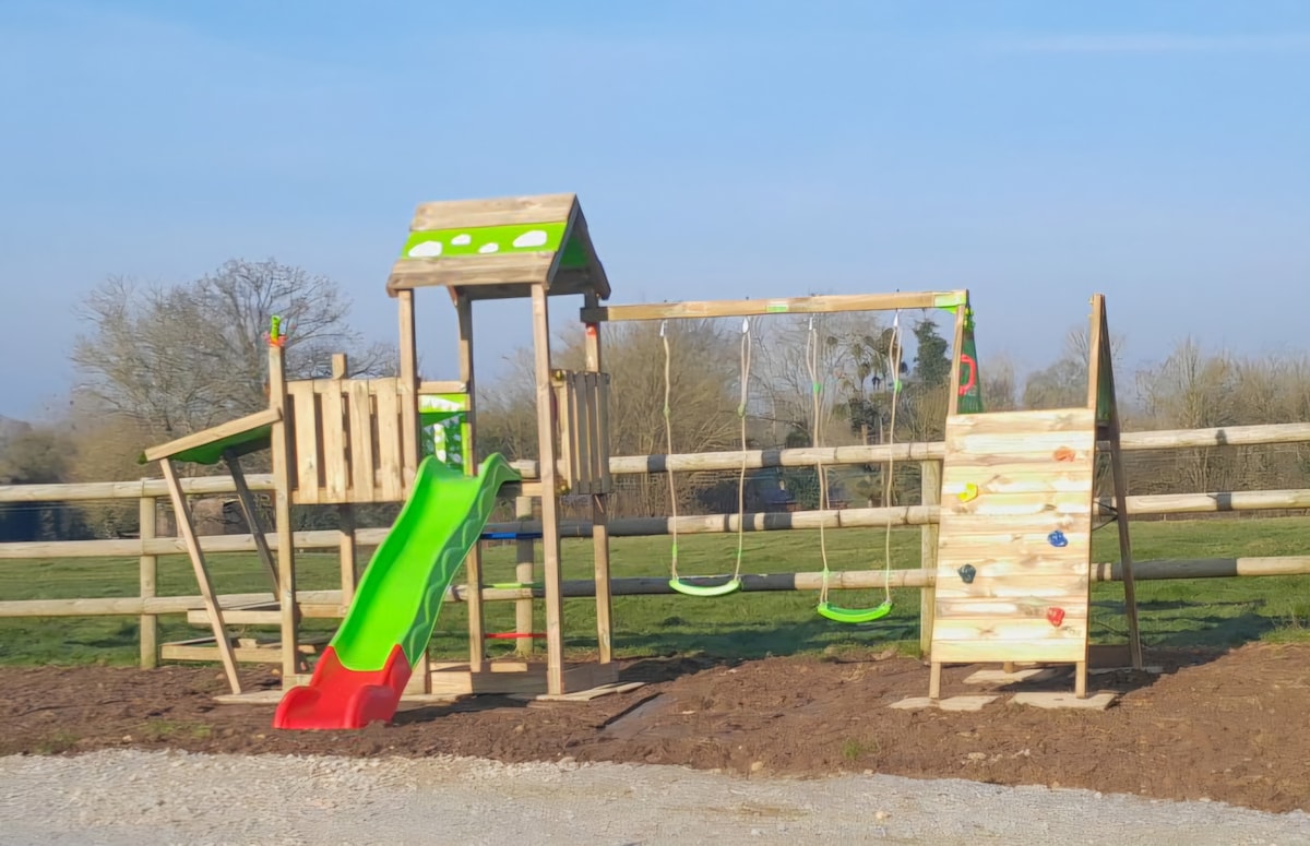 A children’s play area is depicted, featuring a slide in vibrant red and green, swings hanging from a sturdy frame, and a climbing wall with colorful grips. The setup is surrounded by a wooden fence and open grassland, providing a safe and welcoming environment for play.